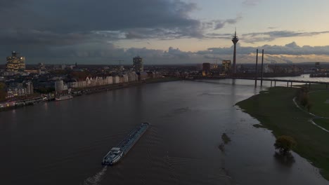 barge sails up flooded rhine river at dusk, dusseldorf, drone tracking shot