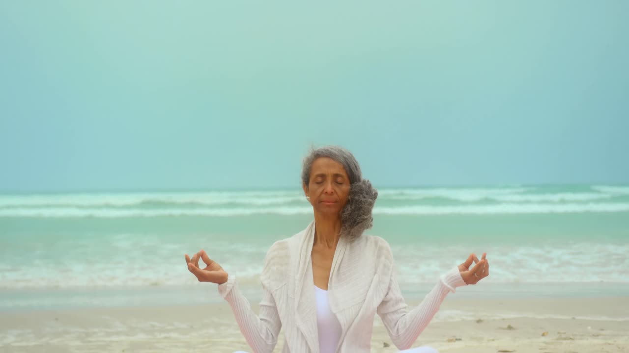 Front view of active senior African American woman doing yoga on exercise mat at the beach 4k