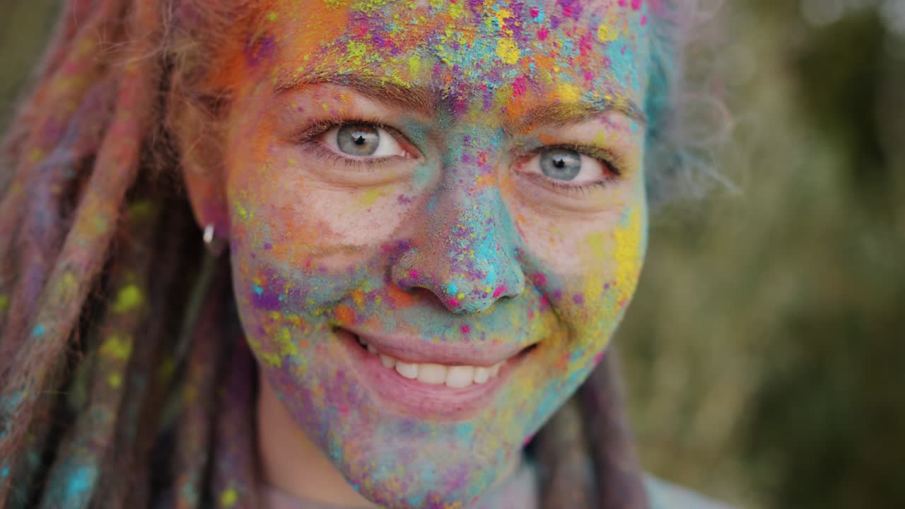 Slow motion portrait of cute girl with colored face and dreadlocks smiling enjoying Holi party