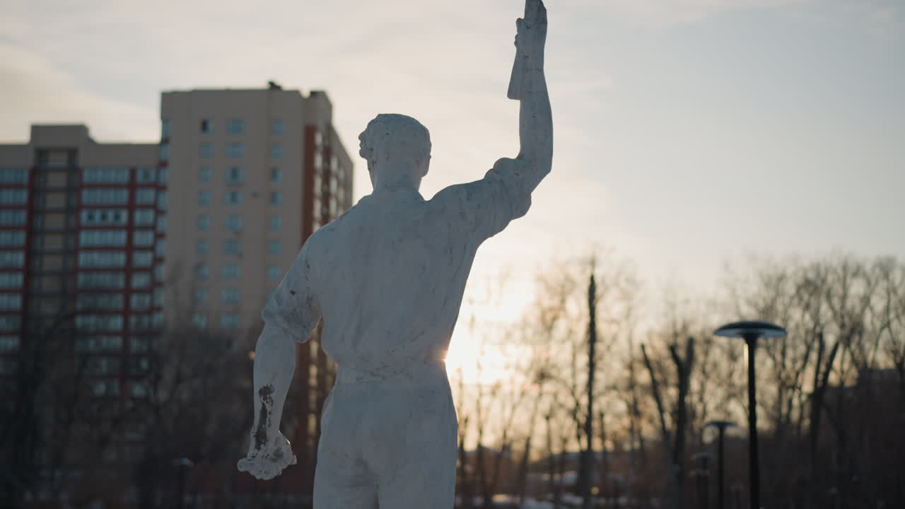 Rear view of sculpture with hand raised against winter sunset glow as person in winter outfit walks past snowy pathway, with bare trees, high rise building, and green lens flare in soft background