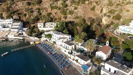 drone view in greece flying over blue sea in loutro small white house town and small boats next to a hill on a sunny day