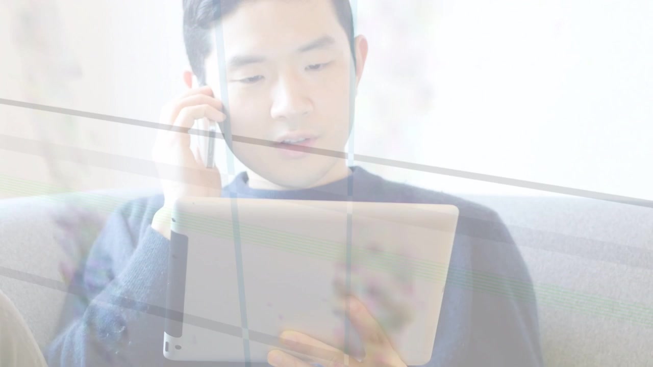 ビジネス通話に応対する男性プロフェッショナルが、タブレットを操作しながら耳を傾ける。明るい窓にはブラインドがかかっている。