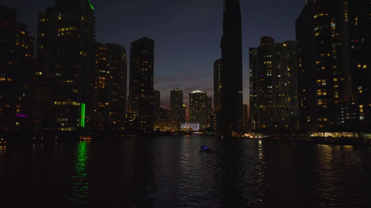 Low angle view of tall buildings with lighted windows around river. Forwards fly above water surface at night. Miami, USA