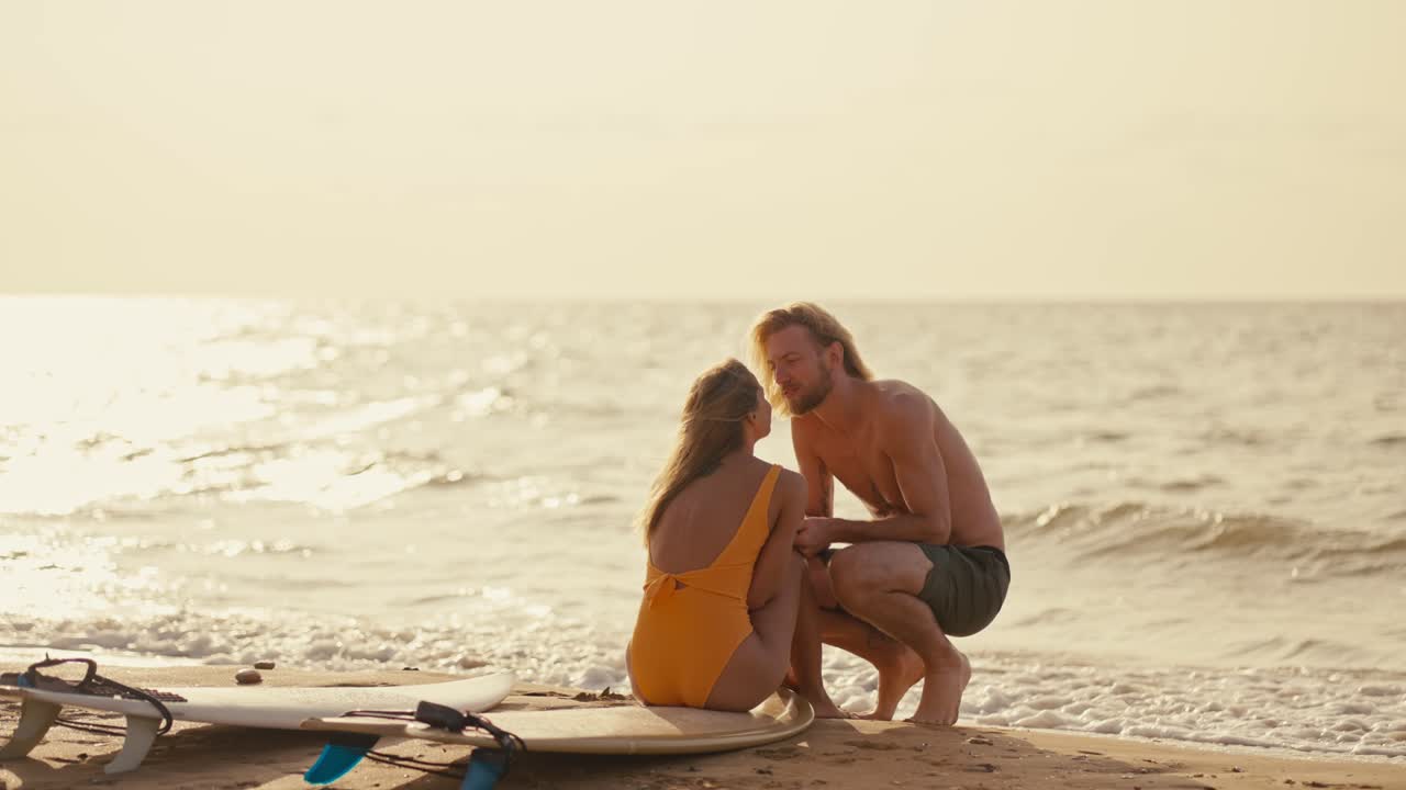 Happy couple on the seashore: a blond guy sits near his blonde girlfriend in an orange swimsuit and kisses her on the seashore in the morning at Sunrise