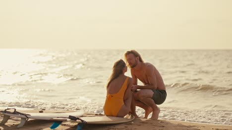 Happy-couple-on-the-seashore:-a-blond-guy-sits-near-his-blonde-girlfriend-in-an-orange-swimsuit-and-kisses-her-on-the-seashore-in-the-morning-at-Sunrise