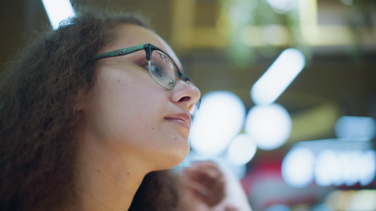 Close-up profile of woman wearing glasses, adjusting hair with serene expression, soft bokeh lights and blurred background providing gentle ambiance, highlighting a reflective moment