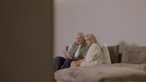 senior couple sitting on sofa at home using digital tablet