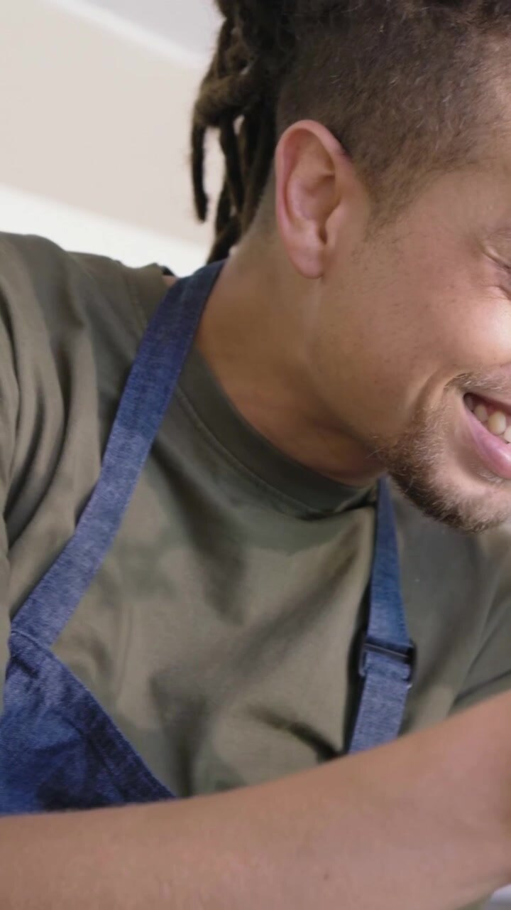 Vertical video: Noticing seedlings, man in olive shirt, blue apron checking pot, working at counter