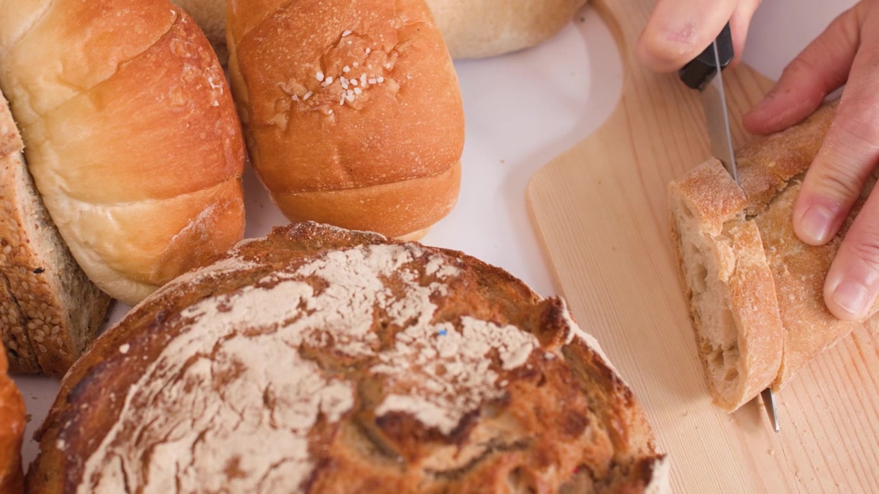 Close-up overhead shot of hands slicing bread on a wooden board with various bakery items.