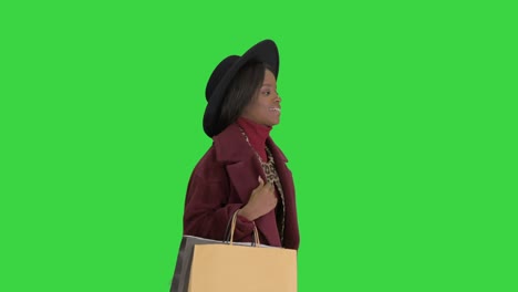 sonriente chica de moda afroamericana en abrigo y sombrero negro posando con bolsas de compras en una pantalla verde, llave de croma