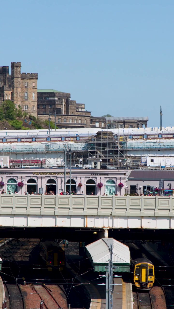 Gulfrontet tog nærmer sig travle jernbaneperroner i klart dagslys, med historiske Edinburgh-bygninger i baggrunden