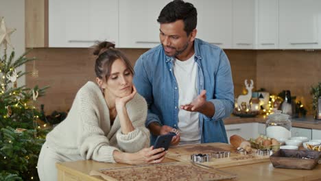 57 latin man making gingerbread cookies and caucasian woman using phone next to him.