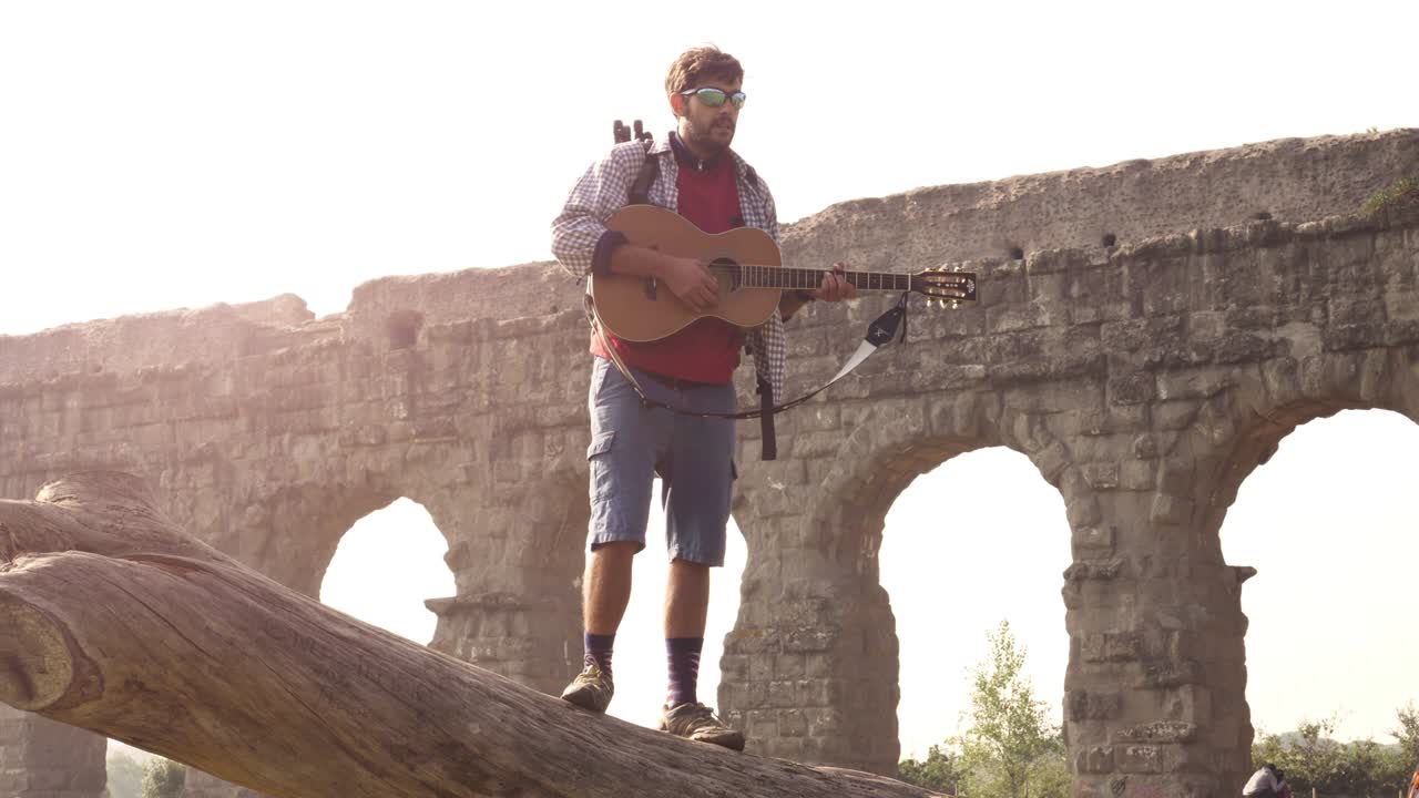 joven aventurero viajero de pie en la parte superior de un tronco de tronco tocando la guitarra cantando frente a las antiguas ruinas del acueducto romano en el parque degli aquedotti en roma al amanecer en cámara lenta