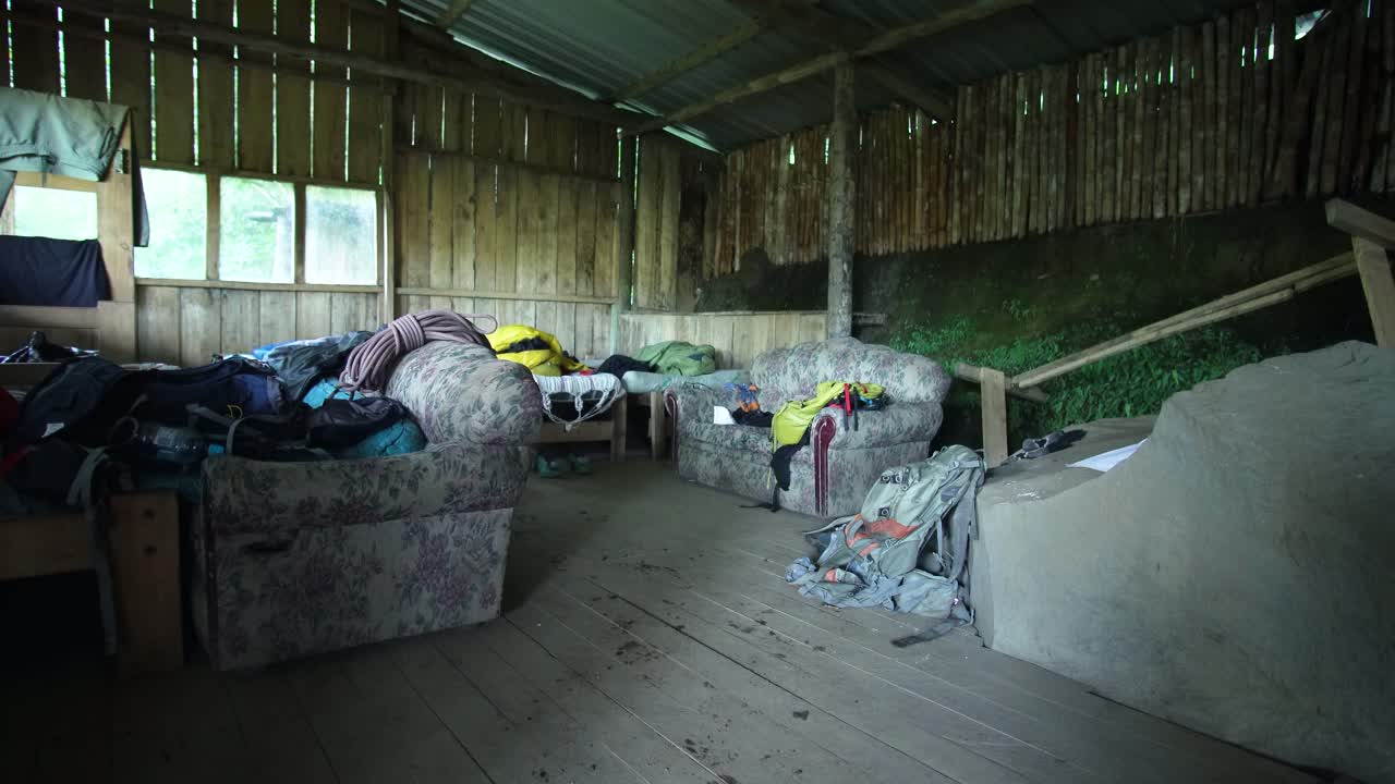 Static shot of a rustic mountain shelter showing old sofas, wooden walls and scattered climbing gear, creating a rugged expedition atmosphere