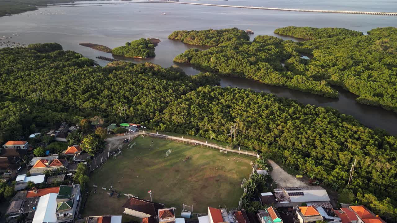 Scenic drone footage captures Bali football stadium area with players, tropical environment, nearby coastal wetlands, and surrounding red rooftops, offering a unique perspective of island recreation