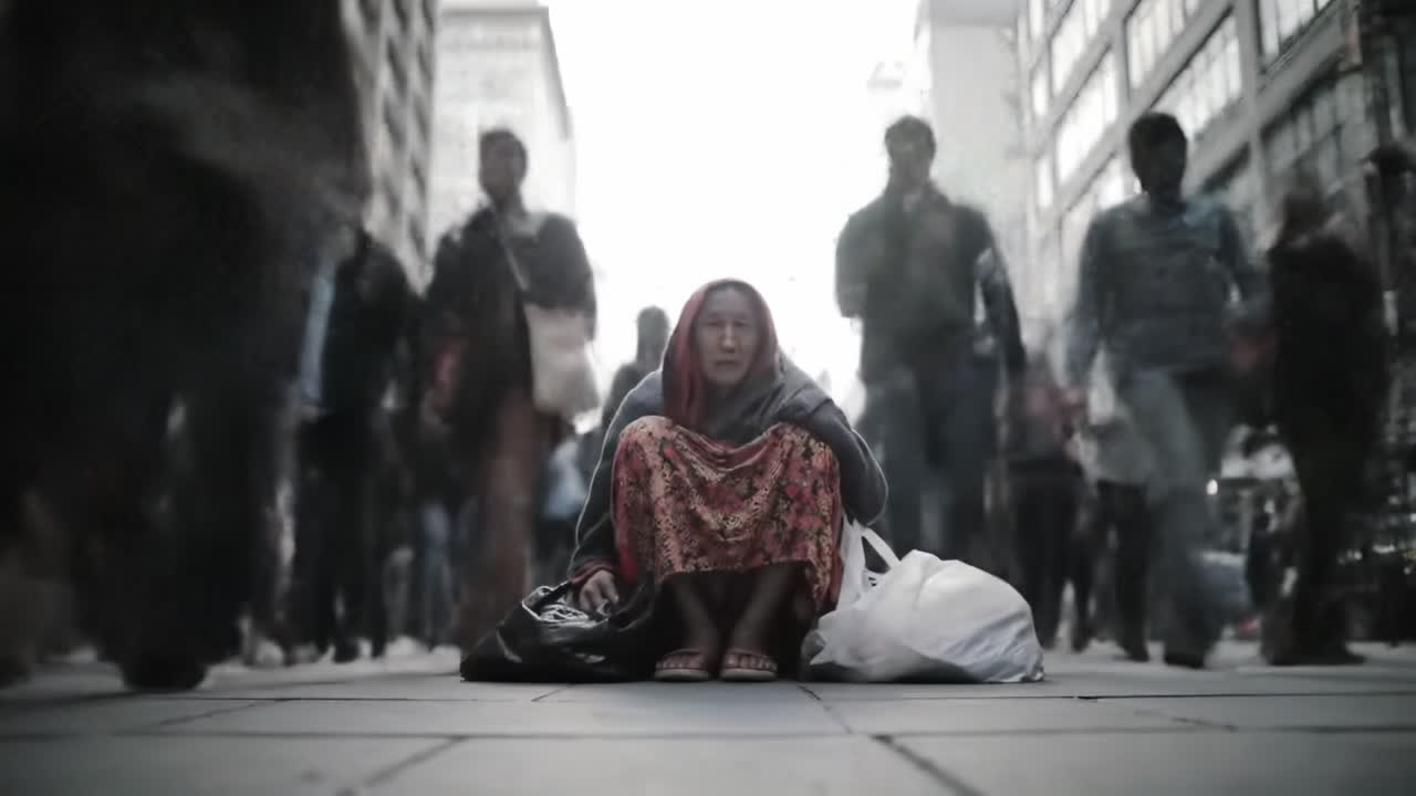 A homeless individual sits on the sidewalk surrounded by a crowd of pedestrians in a busy city.