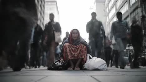 a homeless individual sits on the sidewalk surrounded by a crowd of pedestrians in a busy city.