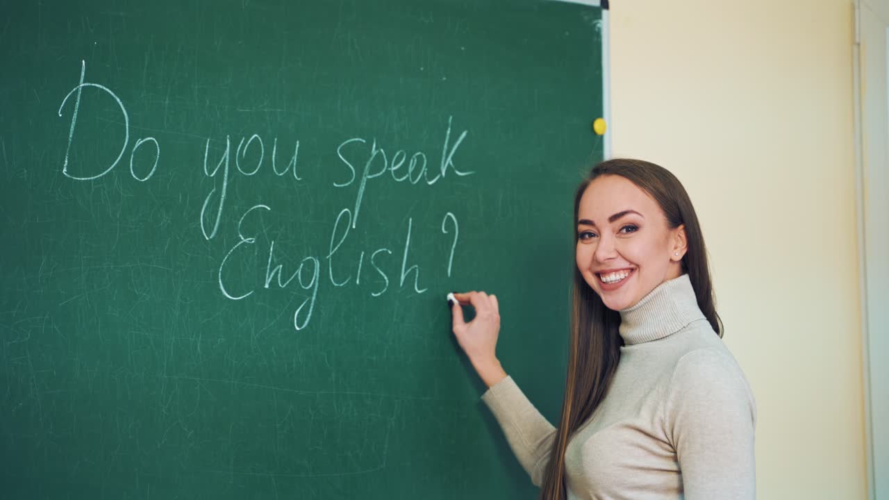 Young teacher writes on a green board with a chalk. Attractive teacher of English puts the question tag on the blackboard and smiles to camera in the classroom.