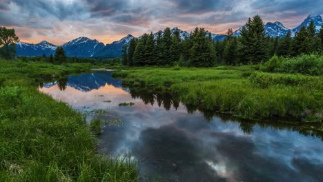 Hermoso-Lapso-De-Tiempo-De-Puesta-De-Sol-Rosa-Y-Naranja-En-El-Lago-Reflectante-Espejo-Con-Montañas-Cubiertas-De-Nieve-Y-Nubes-Gruesas-Y-Esponjosas-Que-Fluyen-En-El-Parque-Nacional-Grand-Teton,-Wyoming,-Estados-Unidos