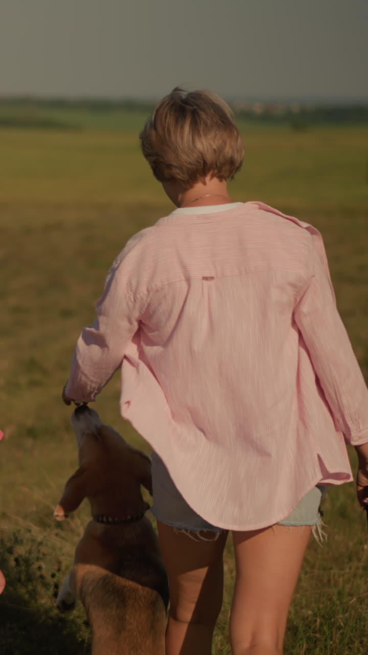 vista trasera de madre e hija caminando estrechamente juntas, con el dueño del perro caminando a su lado mientras el perro se extiende juguetón por su mano, disfrutando de un paseo en un vasto campo abierto rodeado de árboles lejanos