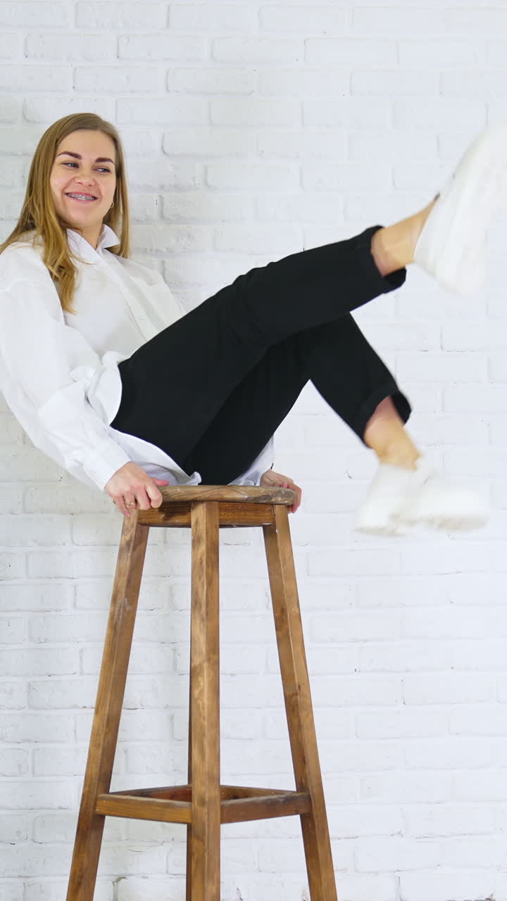 Smiling long-haired lady waving her feet sitting on a high stool. Model demonstrating white loafers with massive soles at white wall backdrop. Vertical video