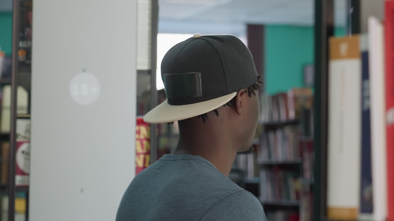 Rear view of man in casual clothing and cap walking through library aisle, surrounded by bookshelves, appearing thoughtful and observant in quiet indoor environment