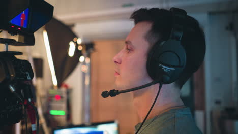 male cameraman wearing professional headset looks attentively at broadcast camera during television studio production, concentrating on filming process with communication gear and soft lighting environment in background