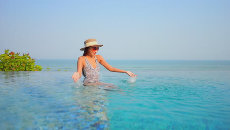 Asian-woman-with-big-sunglasses-and-hat-plays-with-water-in-infinity-pool