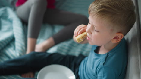 young boy sitting on bed wearing blue shirt eating snack with white plate on lap and partial view of person in leggings beside him capturing casual indoor childhood