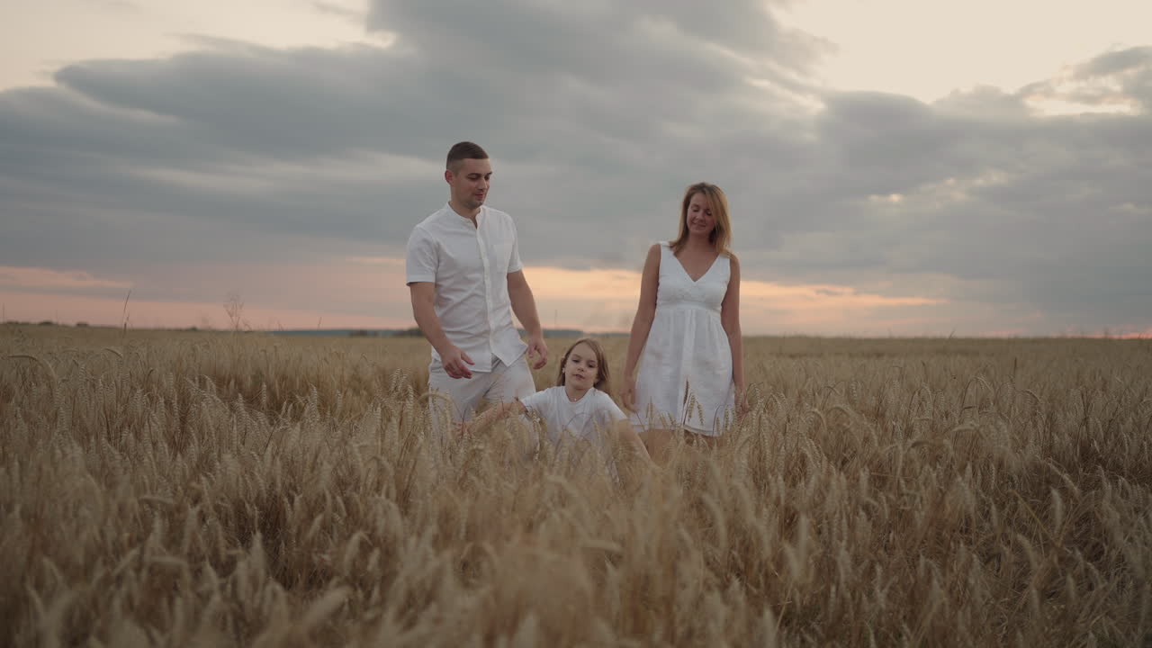 Young couple of parents with girl children holding hands of each other and running through wheat field at sunset. Happy family jogging among barley meadow and enjoying nature together. Slow motion