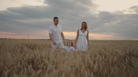 Young-couple-of-parents-with-girl-children-holding-hands-of-each-other-and-running-through-wheat-field-at-sunset.-Happy-family-jogging-among-barley-meadow-and-enjoying-nature-together.-Slow-motion