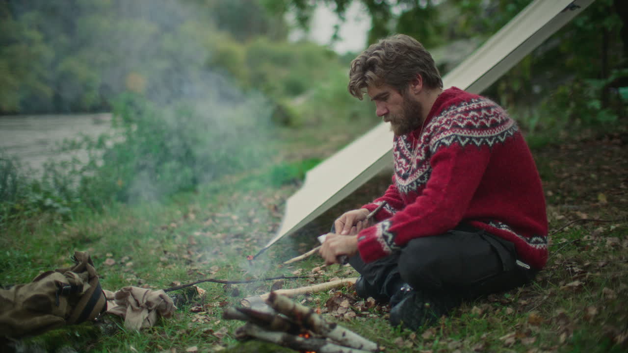 Man Carving Wooden Stick beside Campfire and Tarp Shelter in Forest