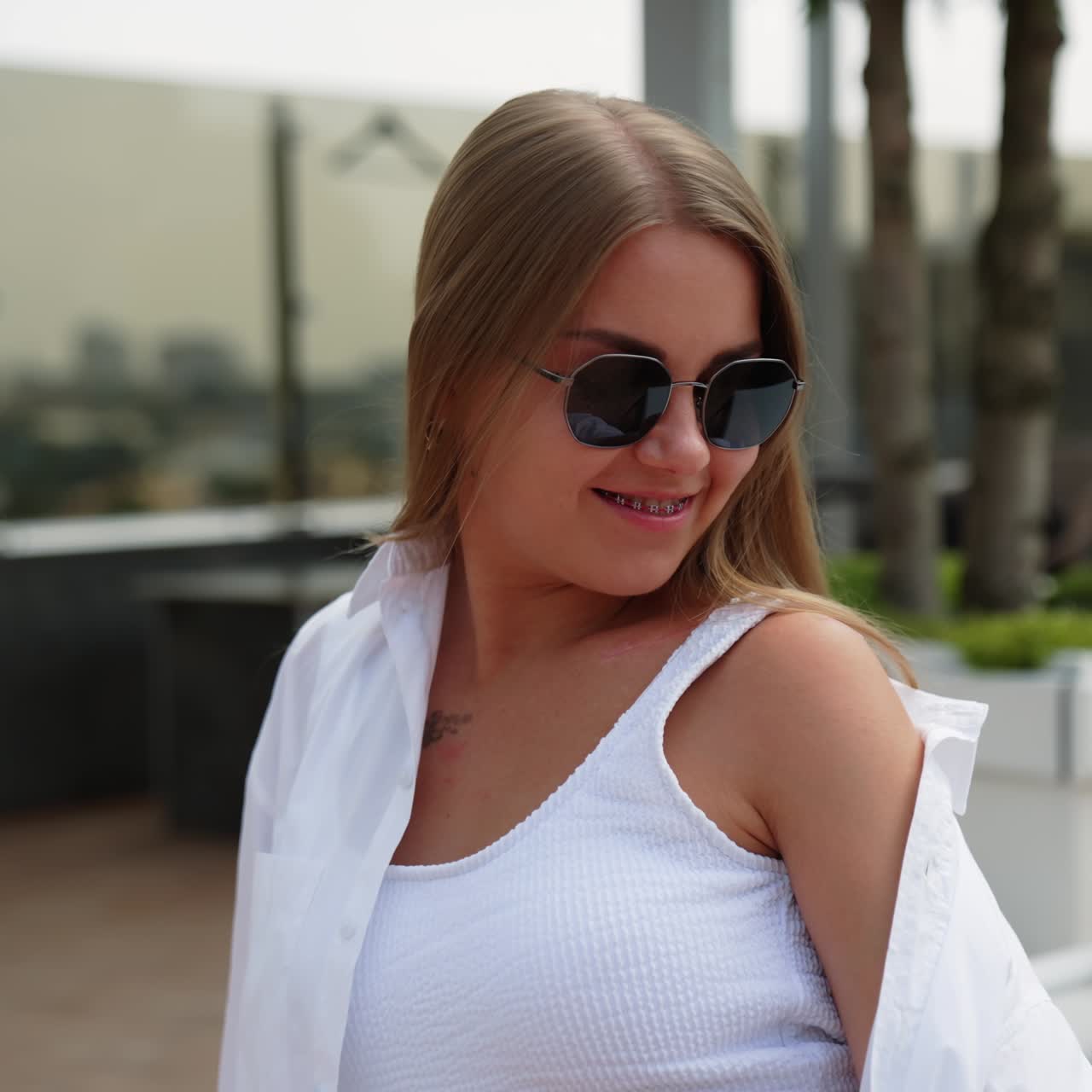 Blonde girl sitting on deckchair in white clothes wearing sunglasses. Smiling woman covers her shoulder from sun