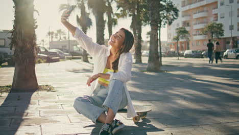 estudiante alegre tomando selfie con el teléfono en el verano. chica despreocupada sentada en longboard