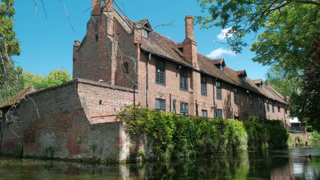 Ampia vista panoramica dell'edificio storico accanto allo stagno a Well Hall Park a Eltham, Londra, con vegetazione lussureggiante, alberi maturi e acqua calma che creano un paesaggio estivo pacifico.