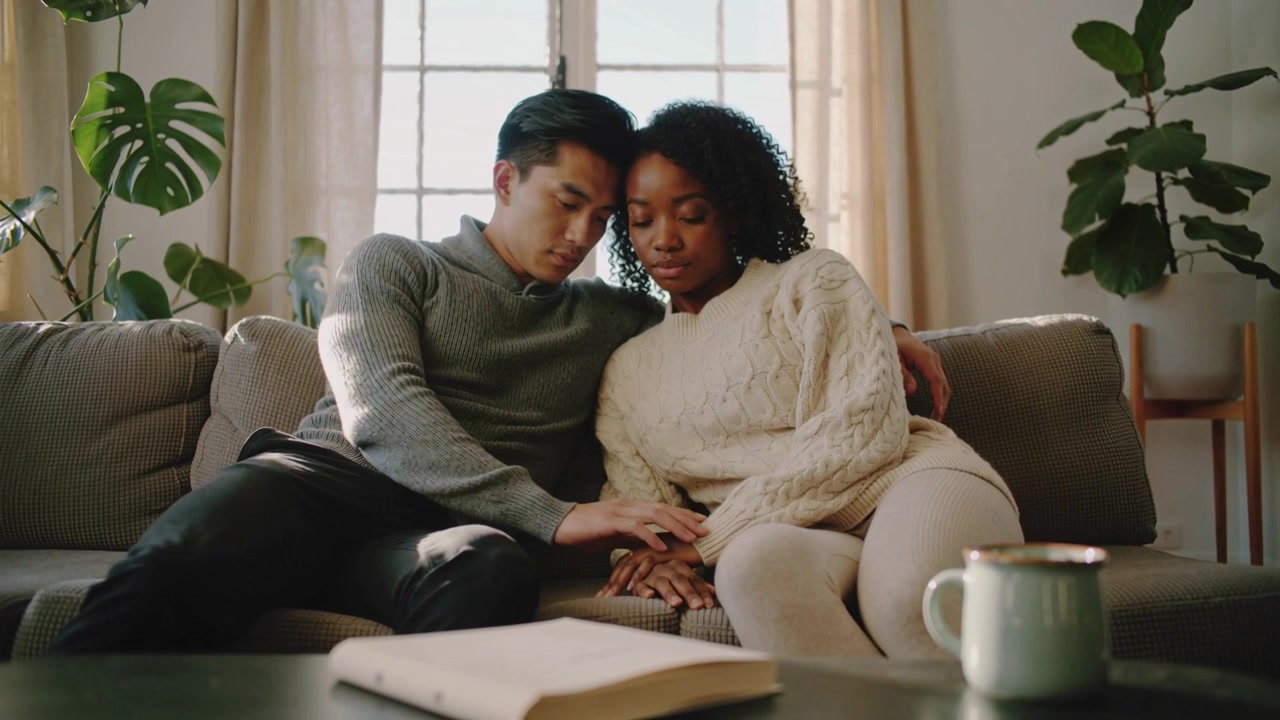 Reaching man in grey sweater touching woman in cream sweater's hand on sofa seeking comfort, mug