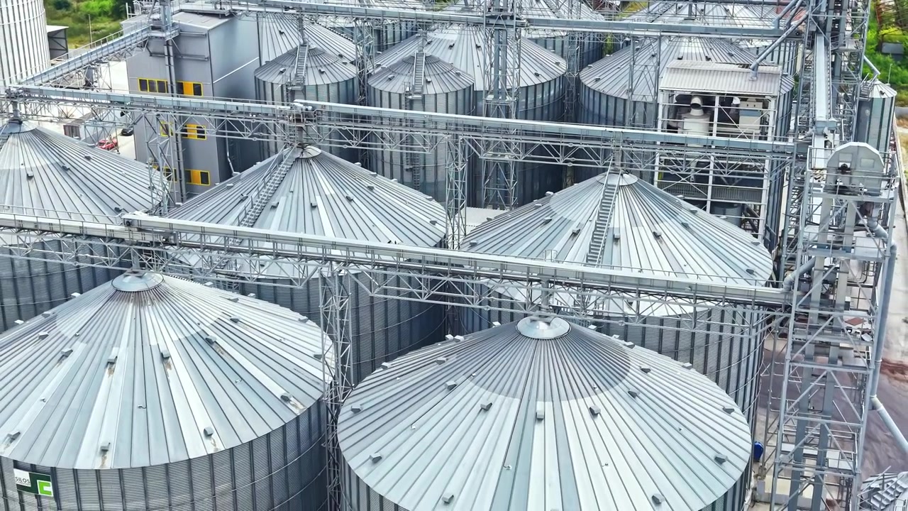 Storage tanks at a grain facility located in a rural area during the day with sunlight highlighting the metal structures and surrounding landscape