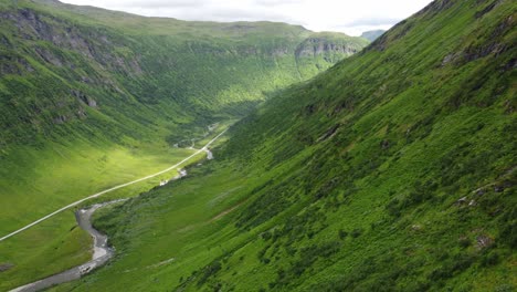 Deep-lush-pristine-valley-of-Kvassdalen-leading-to-Myrkdalen-close-to-Vikafjell-Norway---Aerial-following-green-mountainside-downwards-while-looking-at-sunrays-hitting-river-and-road-in-bottom