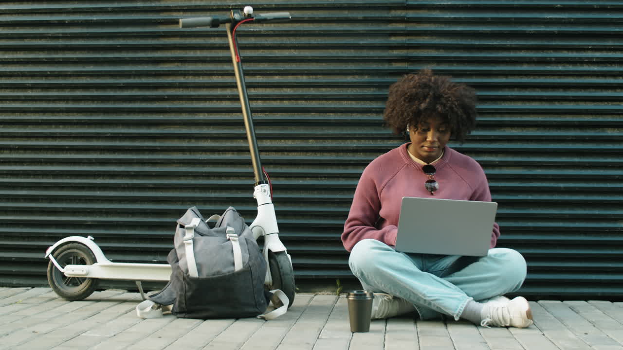mujer afroamericana sentada en la calle en un scooter electrónico y usando una computadora portátil