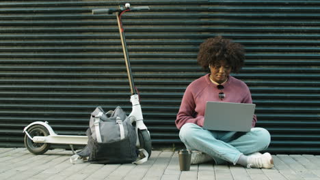 african american woman sitting on street by e-scooter and using laptop