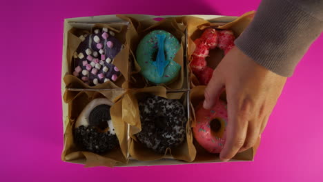 a male hand reaches into a delivery box lined with parchment to take a pink frosted sprinkle donut from a vibrant assortment of six colorful donuts, set against a bright pink background.