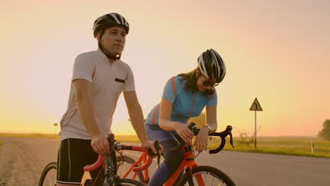 A-man-and-a-woman-in-helmets-with-bicycles-stand-and-talk-at-sunset.-Rest-after-a-bike-ride-on-the-highway.-Track-bikes.-Couple-sports