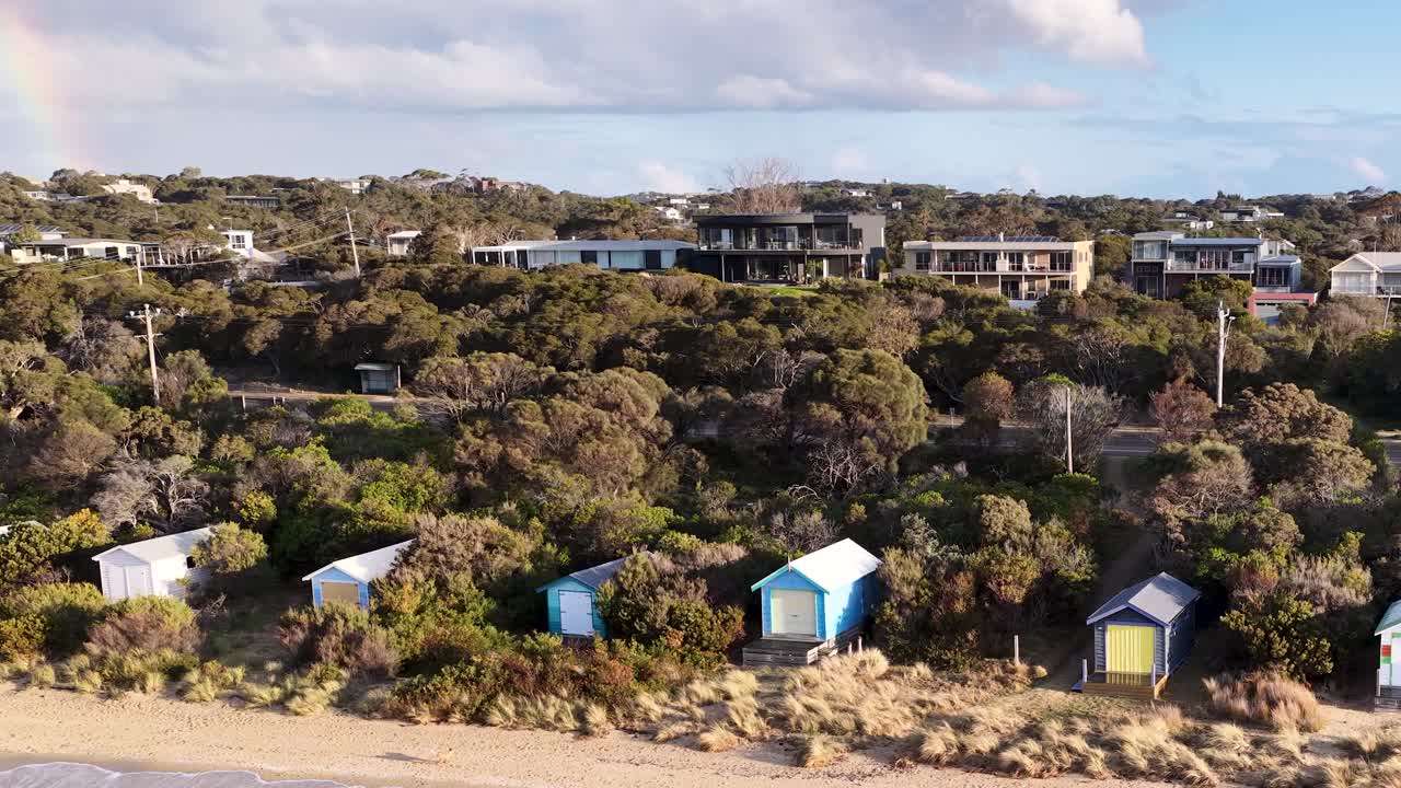 Drone glides above coastal beach huts, bright daylight, revealing lush vegetation and residential backdrop