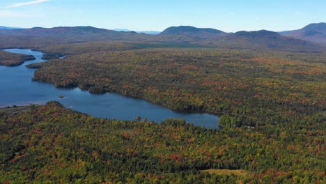 Aerial-drone-shot-tilting-down-over-a-serene-blue-mountain-lake-in-the-forest-along-a-mountain-range-as-summer-ends-and-the-season-changes-to-fall-in-Maine
