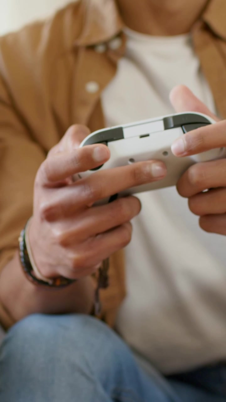 Vertical video: Opening closeup, adult male in tan shirt adjusting controller on couch, gaming