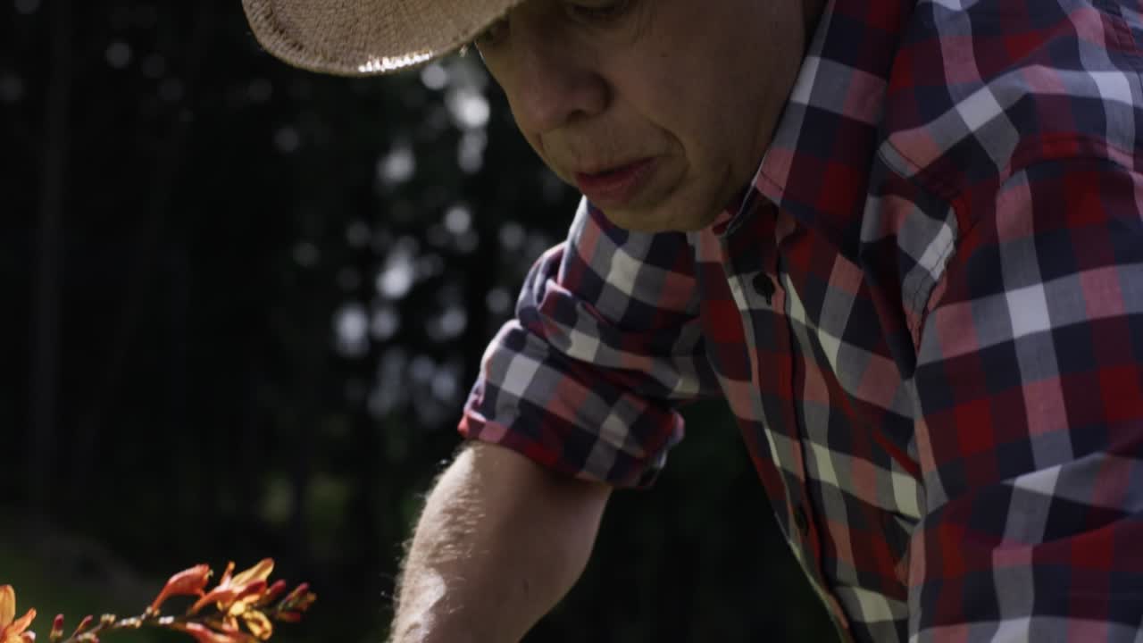 An elderly man in a plaid shirt and a cowboy hat watering the flowers