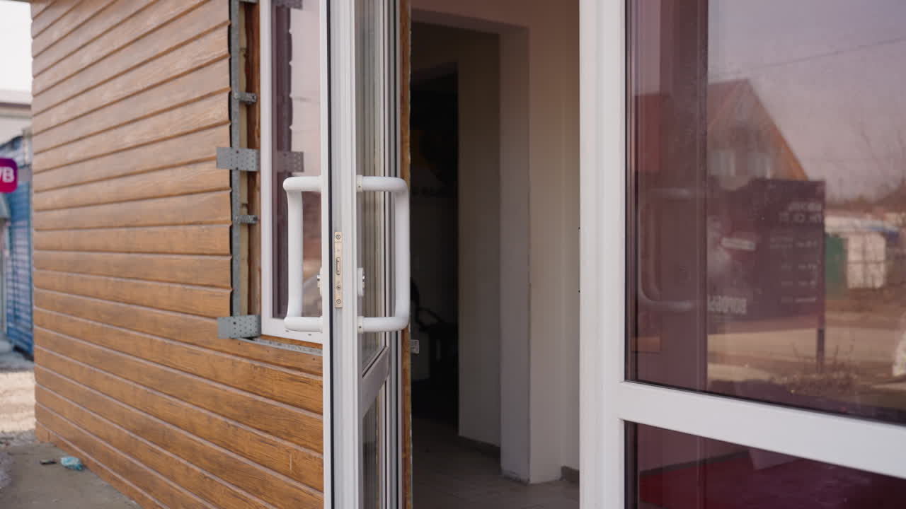 Young man in sunglasses wearing black jacket walks out of a glass door of wooden building, holding handle while stepping outside, another person with leather bag partially visible behind, urban casual lifestyle