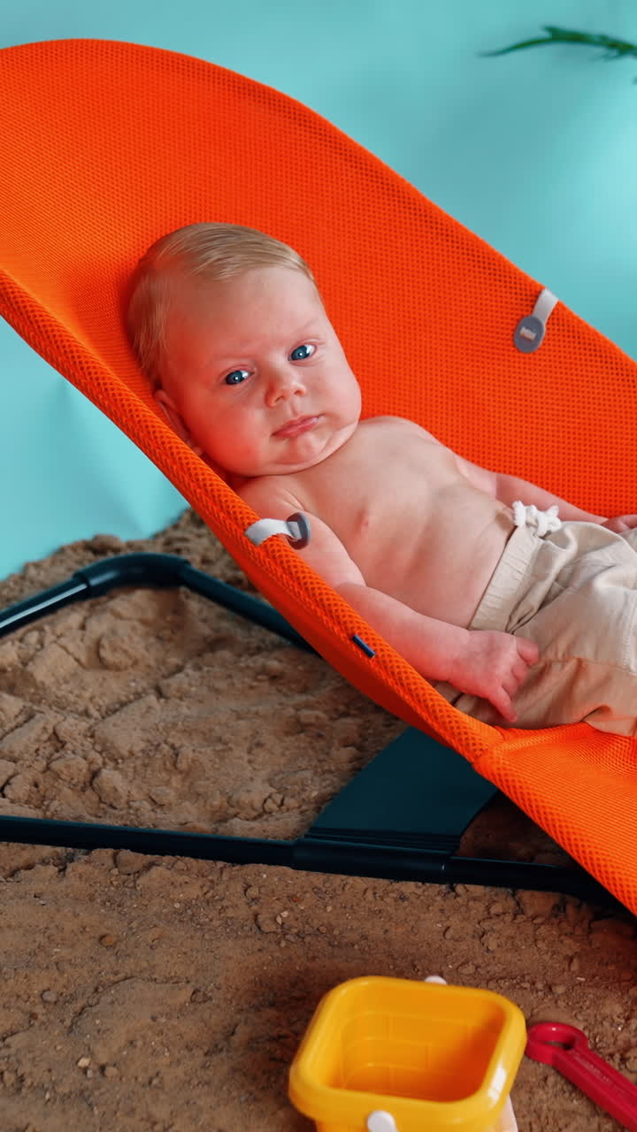 Blond infant boy lies in the orange chair relaxing. Happy vacation on the imitated beach. Side view. Vertical video