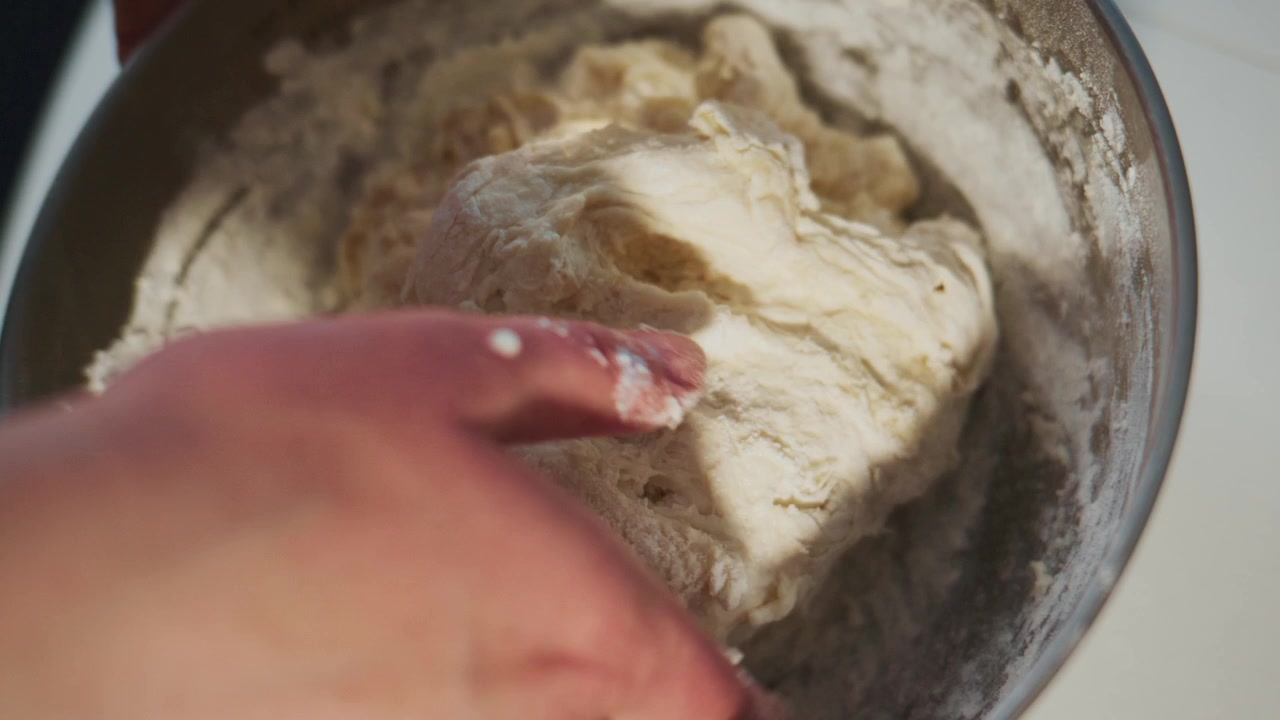 Hand kneads soft dough in metal bowl. Close-up shows flour dusting fingers. Action focuses on tactile food prep. Ideal for recipes, baking tutorials, or culinary branding