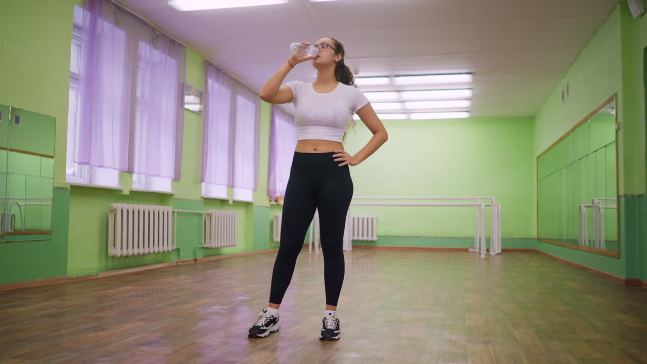 Woman in white and black top standing indoors drinking water from clear bottle calm relaxed posture after workout natural light studio background highlighting hydration recovery wellness fitness